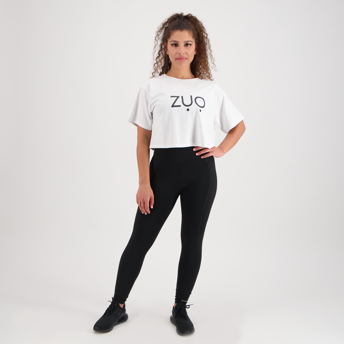View from front of model with curly hair in Keep Up, high waisted black leggings and ZUO Apparel Branded Box Tee. White background. 